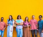 Seven diverse young adults stand side-by-side in front of a vibrant yellow wall, smiling and holding drinks. They appear relaxed and friendly, with a mix of casual outfits and assistive devices visible, symbolising inclusion and community.