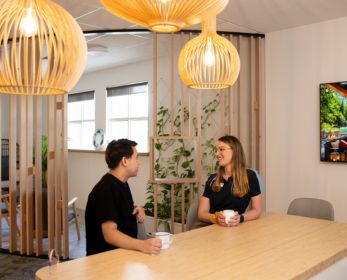 Two students sitting at a wooden table in a warm, modern space, chatting and holding mugs. Pendant lights hang overhead, and indoor plants are visible in the background.