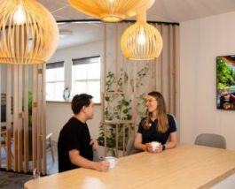 Two students sitting at a wooden table in a warm, modern space, chatting and holding mugs. Pendant lights hang overhead, and indoor plants are visible in the background.
