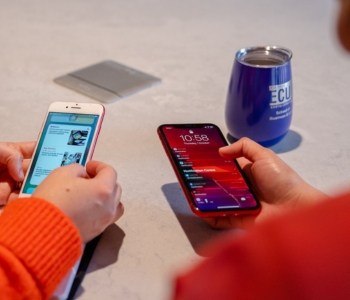 Two people holding smartphones at a table, with an ECU-branded mug in the background.