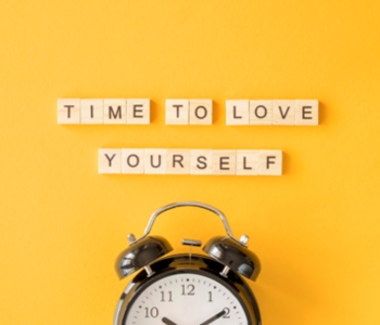 A black alarm clock sits below wooden letter tiles on a bright yellow background. The tiles spell out “TIME TO LOVE YOURSELF,” reinforcing a message of self-care and reflection.