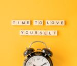 A black alarm clock sits below wooden letter tiles on a bright yellow background. The tiles spell out “TIME TO LOVE YOURSELF,” reinforcing a message of self-care and reflection.