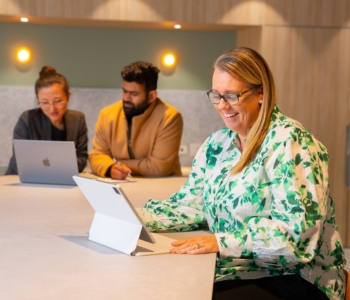 Three ECU students working together on laptops at a study table, with one student smiling while using a tablet.