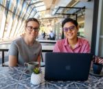 Two students sit at an outdoor café table with coffee and a laptop, smiling toward the camera. The setting is relaxed and open, with natural light streaming through the campus walkway.