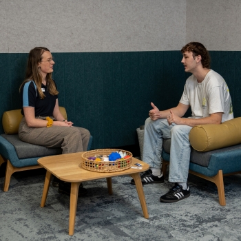 Two students sit across from each other in a relaxed conversation space. One is speaking while the other listens attentively. A low table with sensory items sits between them.