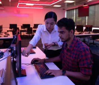Two students working together at a desktop computer in a modern computer lab with ambient red lighting. One is seated and typing while the other points at the screen, offering guidance.