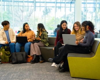 Six ECU students sit on library couches, chatting and working on laptops during a collaborative study session.