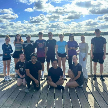 Group of ECU students standing and kneeling on a wooden boardwalk by the water under a cloudy sky, wearing activewear. ECU Active logo in the top left corner.