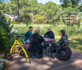 Four students sit together at an outdoor table by a lake on ECU’s campus, including two wheelchair users and an adaptive bicycle, enjoying a conversation in the sunshine.