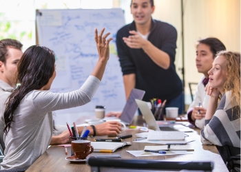 Student raising hand during a collaborative discussion in a focus group setting, with notebooks, laptops, and a whiteboard in the background.