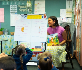 A smiling early childhood teacher sits at the front of a classroom holding the book Zog while young students sit on the floor, engaging in a literacy activity.