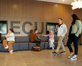 Students chat and walk through the South West ECU lobby, seated and passing in front of a timber feature wall with large “ECU” letters.