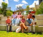 A group of six students sit and chat on tiered outdoor concrete steps under a sunny sky. They’re relaxed, smiling, and enjoying a casual conversation, surrounded by greenery and modern campus buildings.