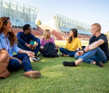 Five students sitting on a grassy area on campus chatting and smiling. 