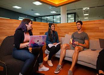 Three students are sitting and chatting in the Library, one of them has a pink laptop.